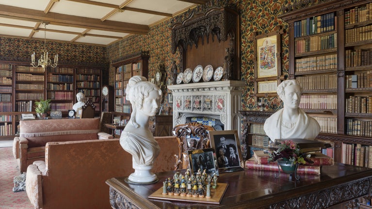 View of the Library, Oxburgh Estate, Norfolk, featuring richly carved tables, sofas, marble busts and variety of objects. The two 19th-century tables may be by Belgian cabinetmaker Malfait, while the marble portrait busts in the foreground depict Mrs Agnes Molyneux Seel and her husband.